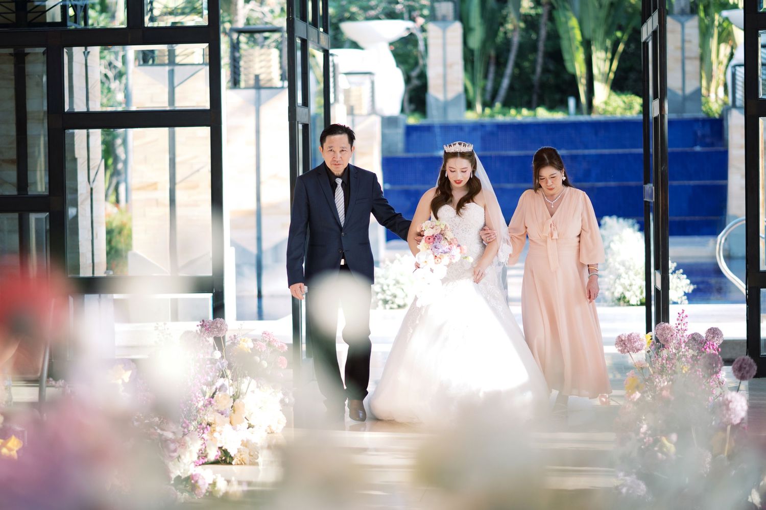 Bride in white gown walks down the aisle with two escorts at an elegant floral wedding ceremony.