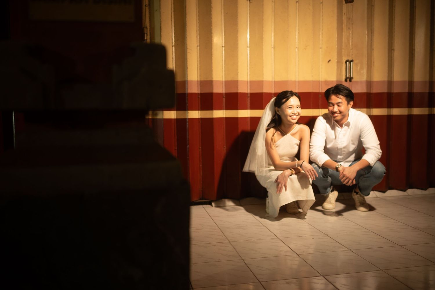 Asian couple in wedding attire smiling and crouching together against a striped wall in warm light.