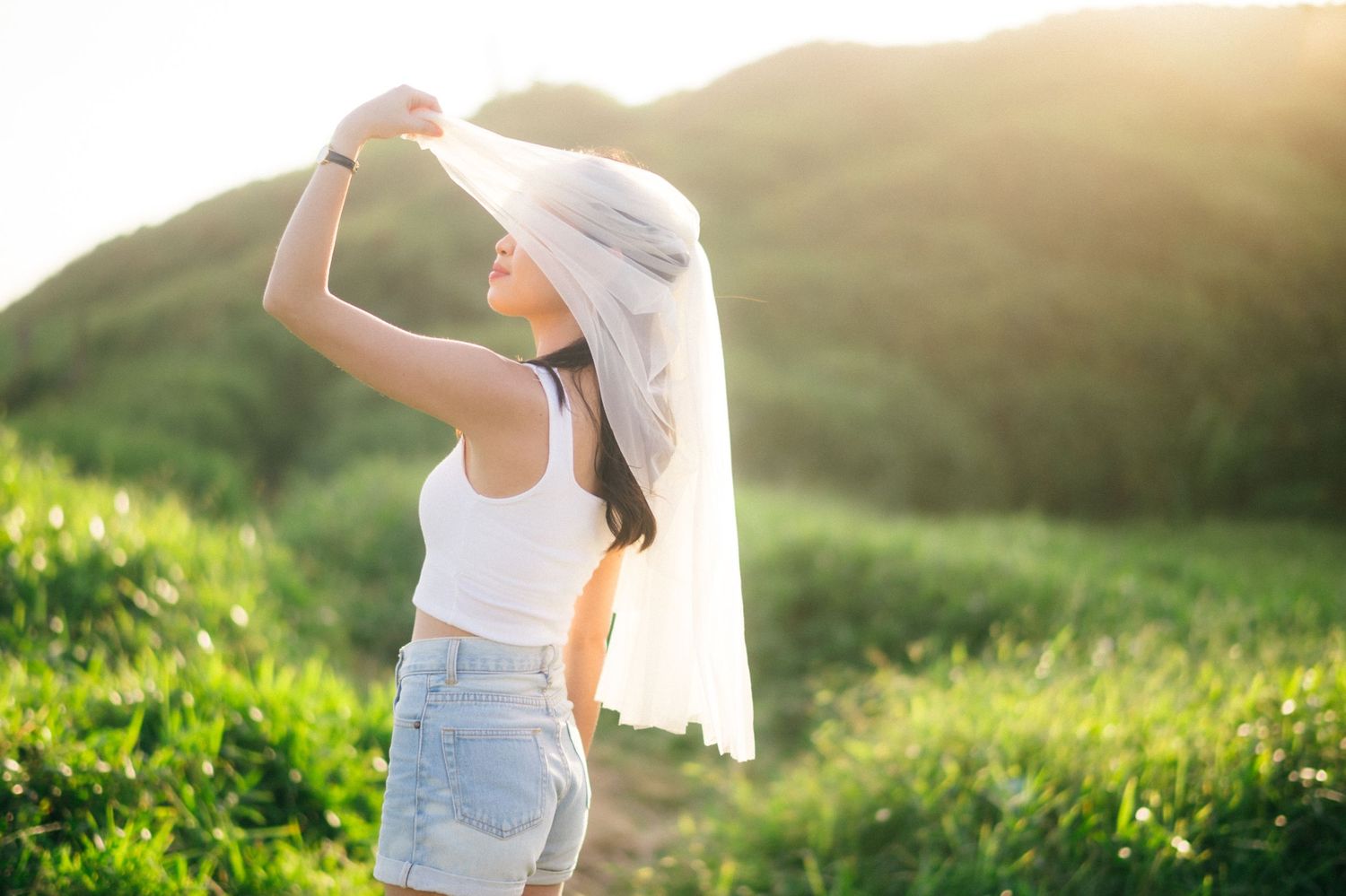Young woman in white top and denim shorts holding a white veil hat outdoors in sunlit green hills.