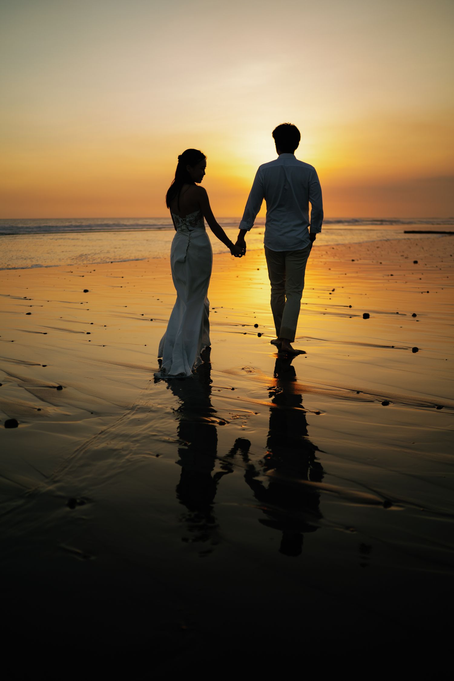 Silhouette of couple holding hands on beach at vibrant orange sunset, reflections on wet sand.