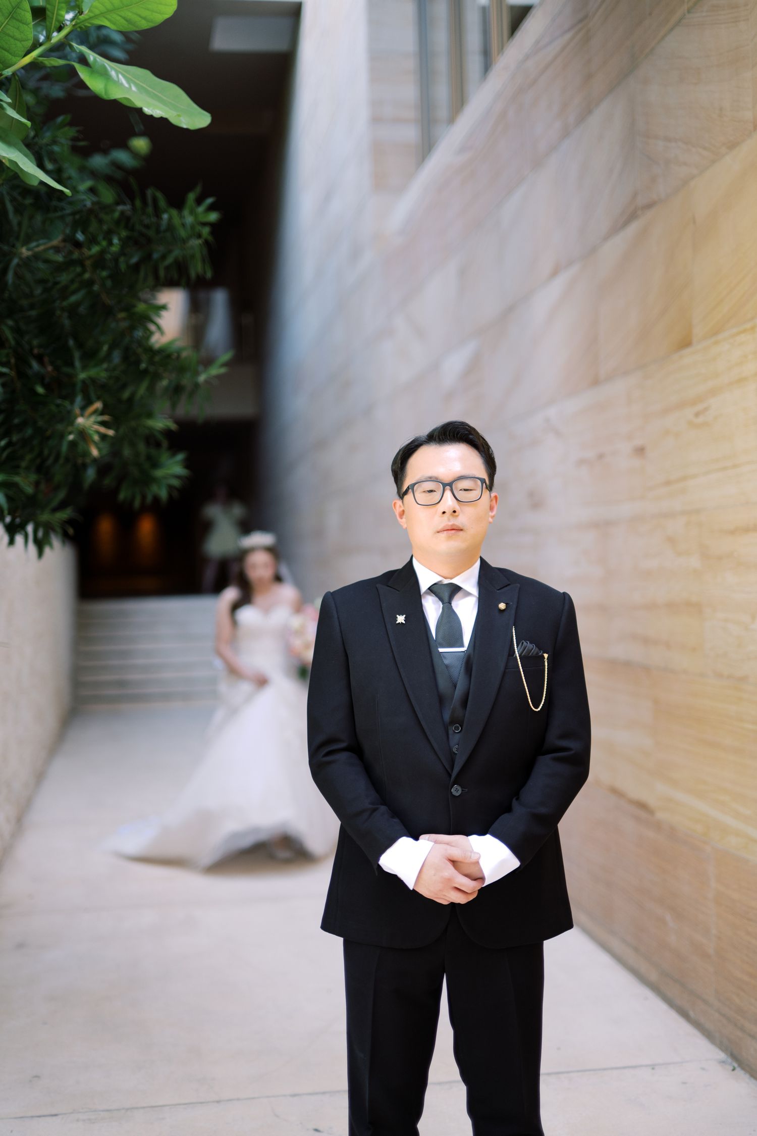 Groom in black tuxedo stands outdoors as bride in white gown approaches in background at wedding.