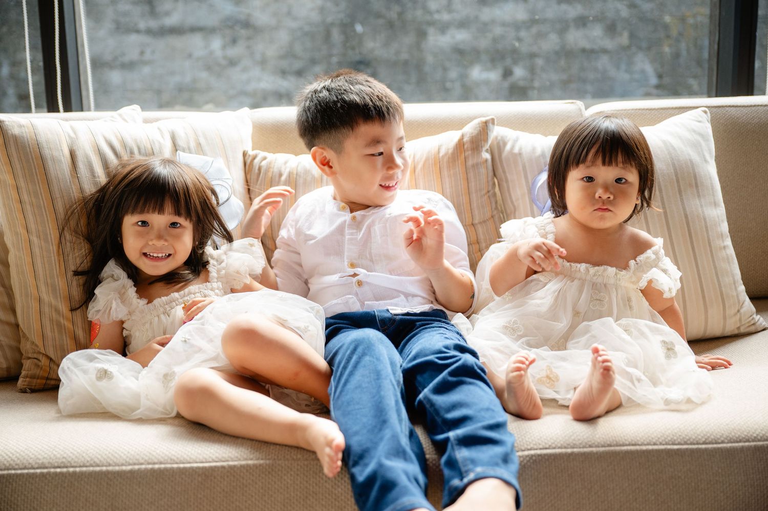Three young children sit together on a beige sofa, two girls in white dresses and a boy in blue jeans, smiling happily.
