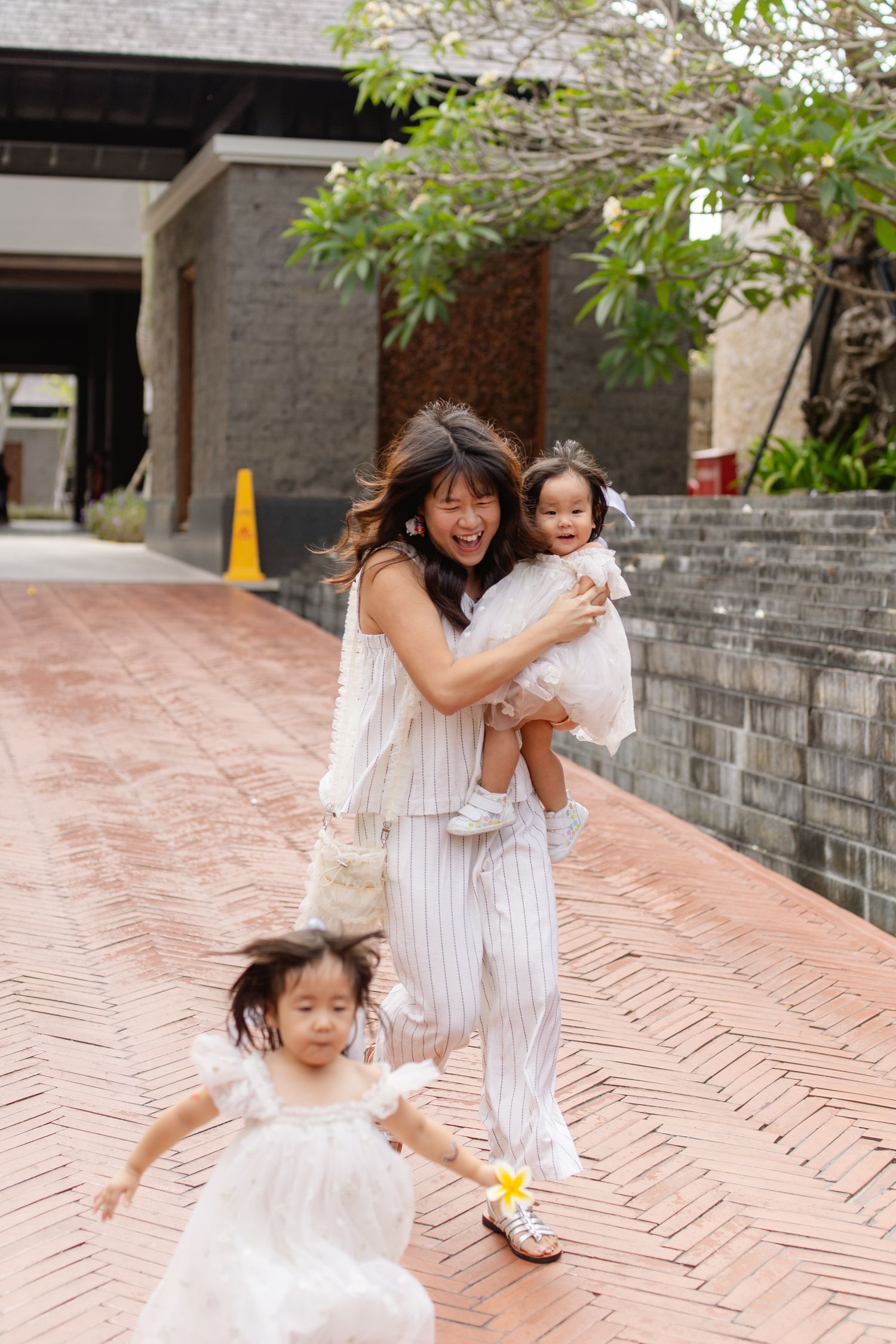 A smiling Asian woman holds a baby girl while another young girl in a white dress runs ahead on a brick pathway.