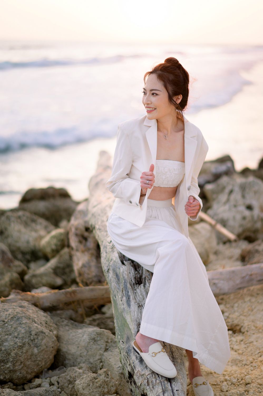 Woman in white blazer and wide-leg pants sitting on driftwood at a rocky beach during golden hour sunset.