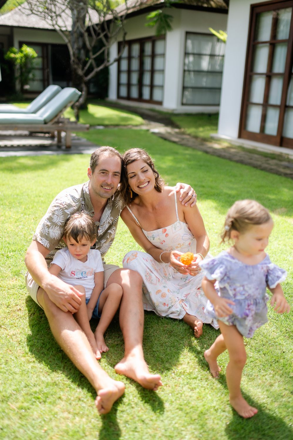 A happy family with two young girls sits on green grass in front of a tropical villa on a sunny day.