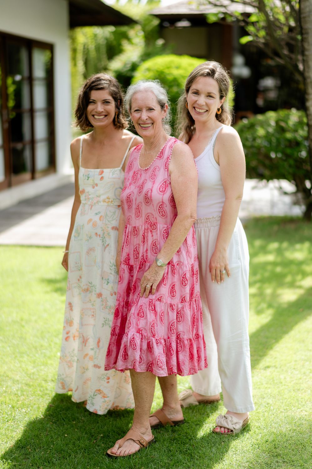 Three smiling women in summer dresses posing together outdoors on green grass near a house.