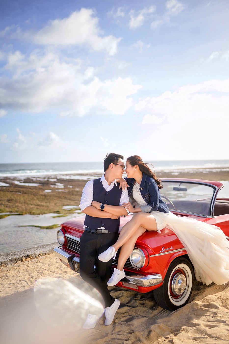 Couple in wedding attire sitting on a classic red convertible car on a sandy beach with ocean backdrop.