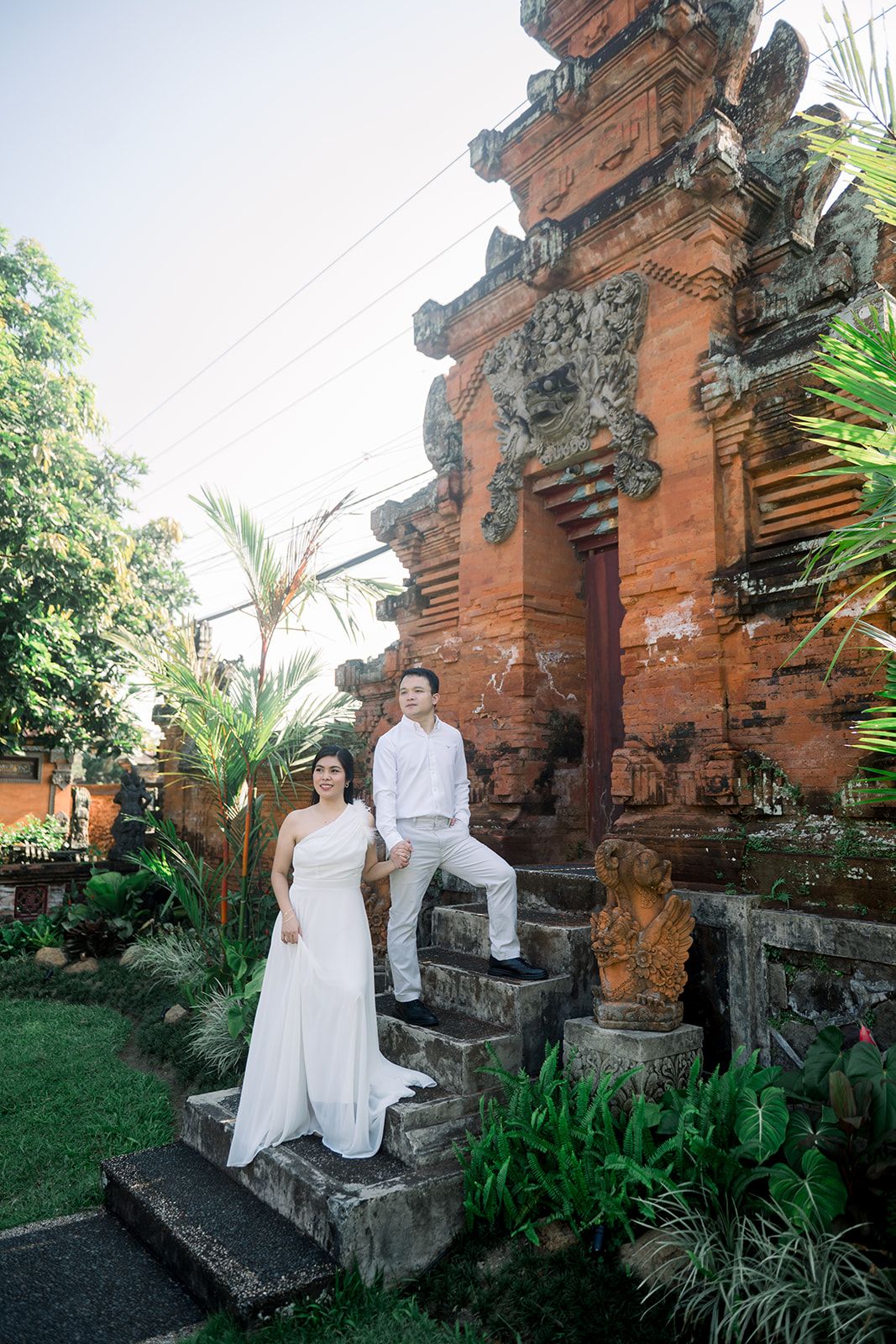 Couple in white attire posing on stone steps of a traditional Balinese temple surrounded by lush tropical greenery.