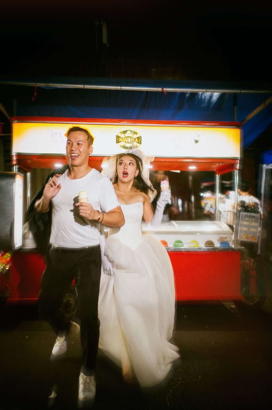 Bride in white gown and groom in white tee laugh excitedly at night beside a colorful food cart.