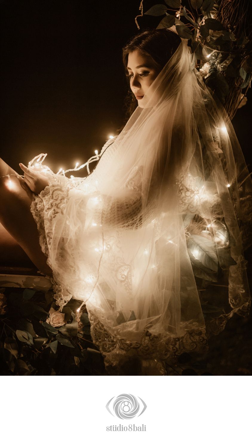 Bride in lace wedding dress and veil surrounded by glowing fairy lights in a dark, romantic outdoor setting.
