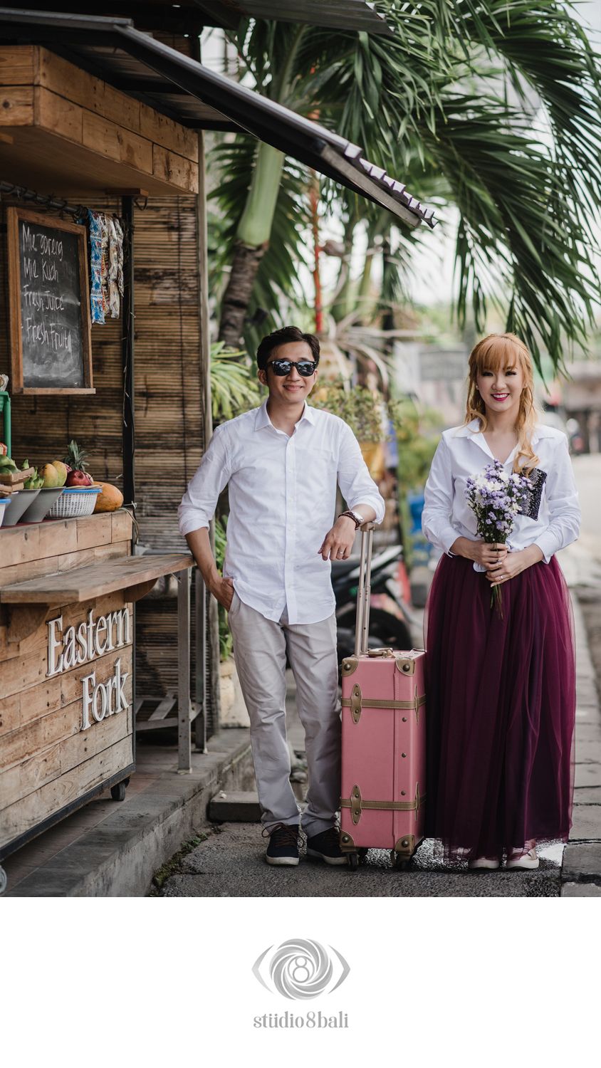 Couple posing outside Eastern Fork cafe in Bali, woman holding purple flowers beside a pink vintage suitcase.