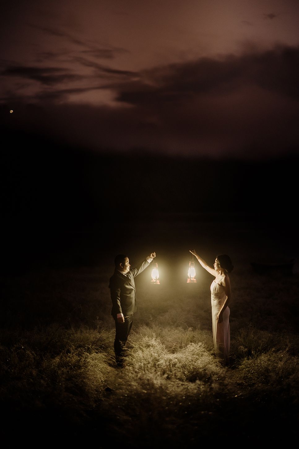 Couple holding glowing lanterns facing each other in a dark field at night, creating a romantic and magical atmosphere.