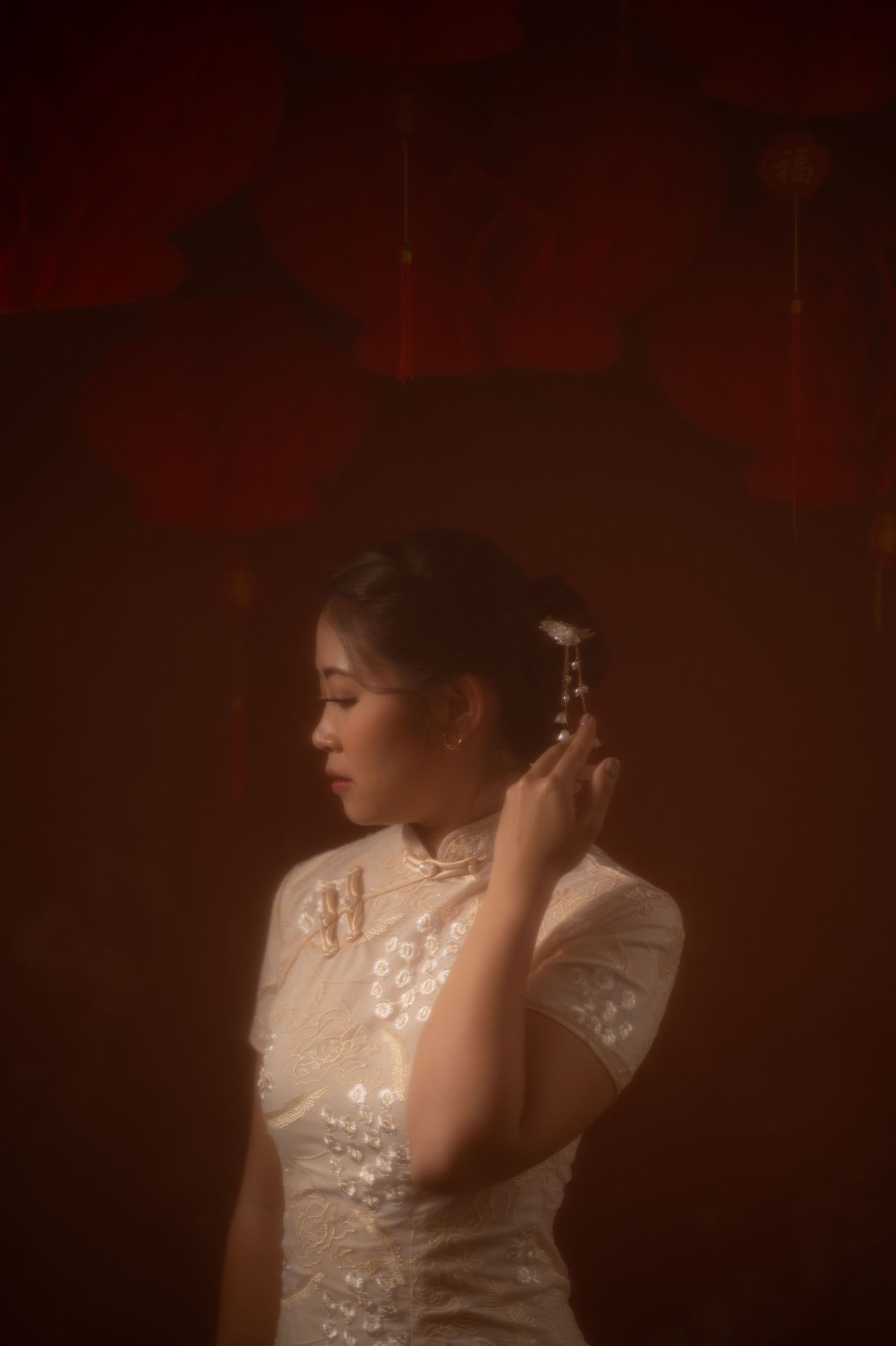 A bride in a white lace qipao adjusts her floral hair accessory against a moody red backdrop.