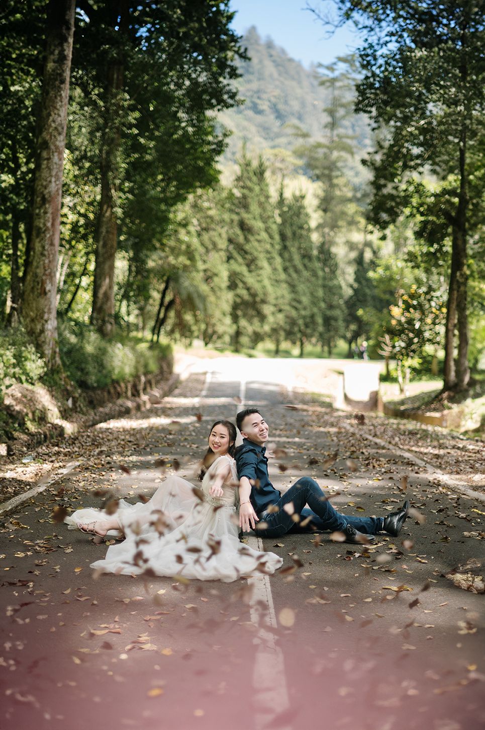 Couple sitting back-to-back on a tree-lined path in a forest, woman in white dress, man in dark jeans.