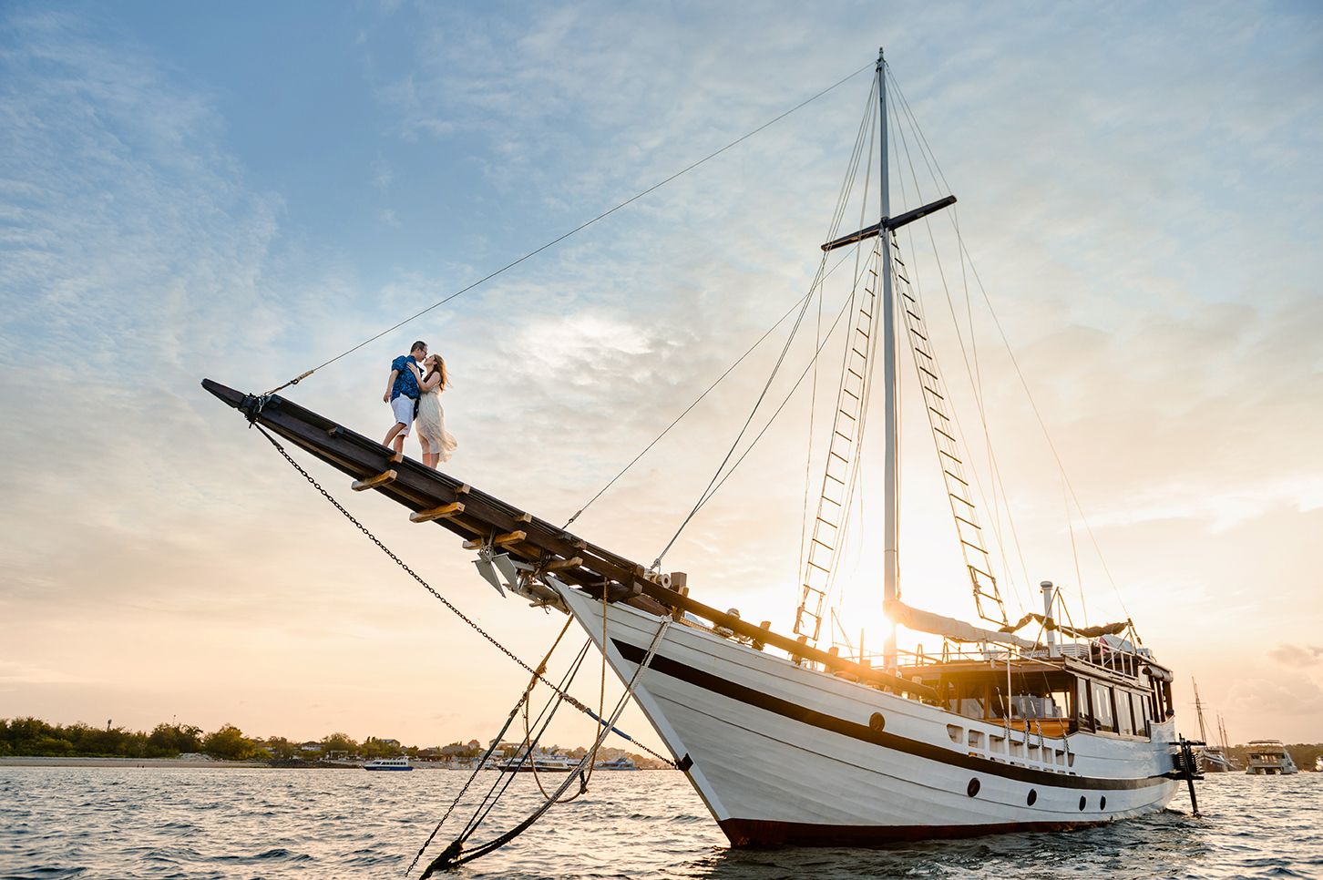 Couple sharing a romantic kiss on the bow of a white sailboat at sunset over calm waters.