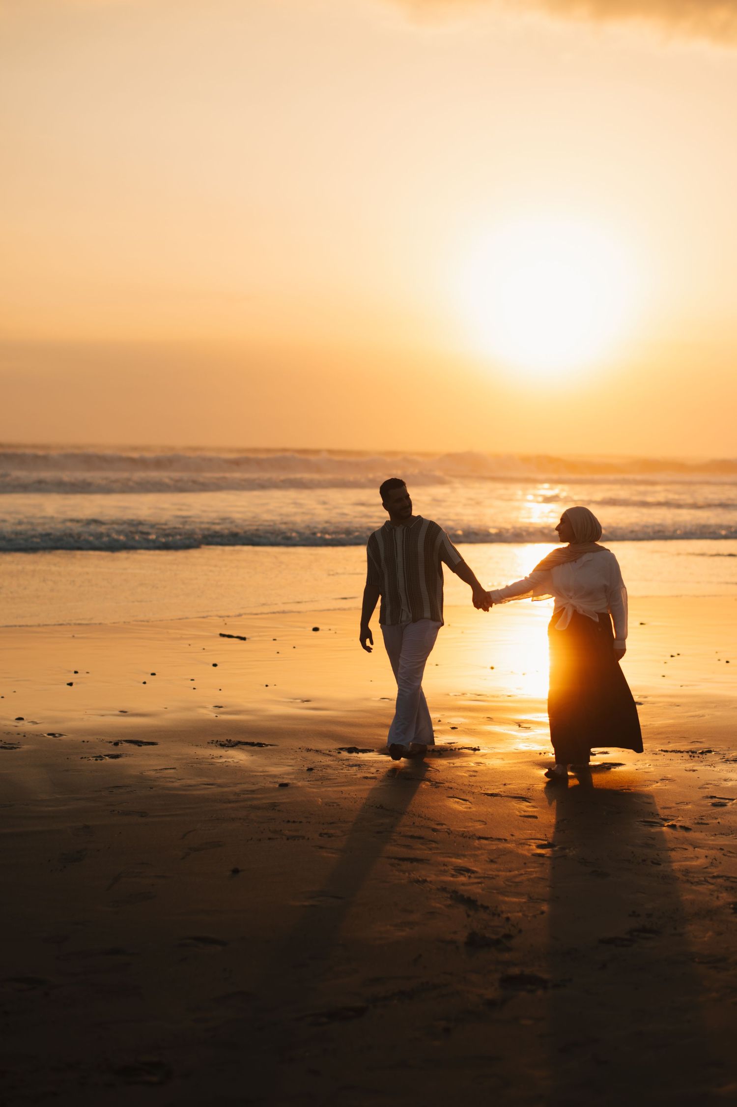 Couple holding hands walking on a sandy beach at golden sunset with ocean waves in the background.