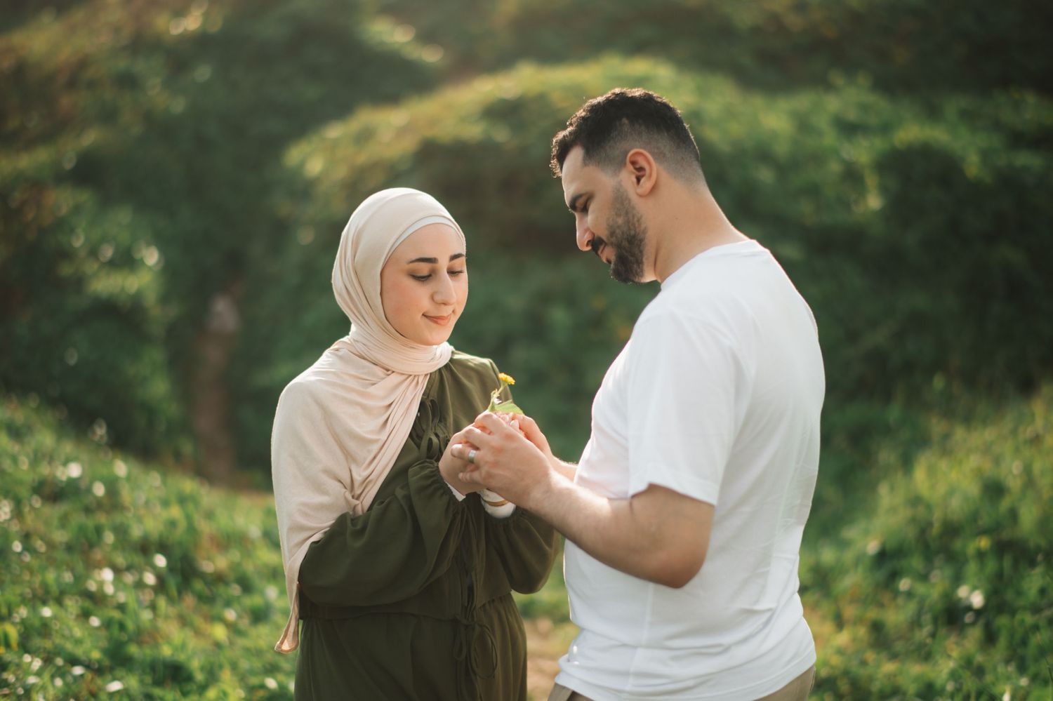 A couple in a green field, woman in hijab and man in white shirt sharing a tender moment outdoors.
