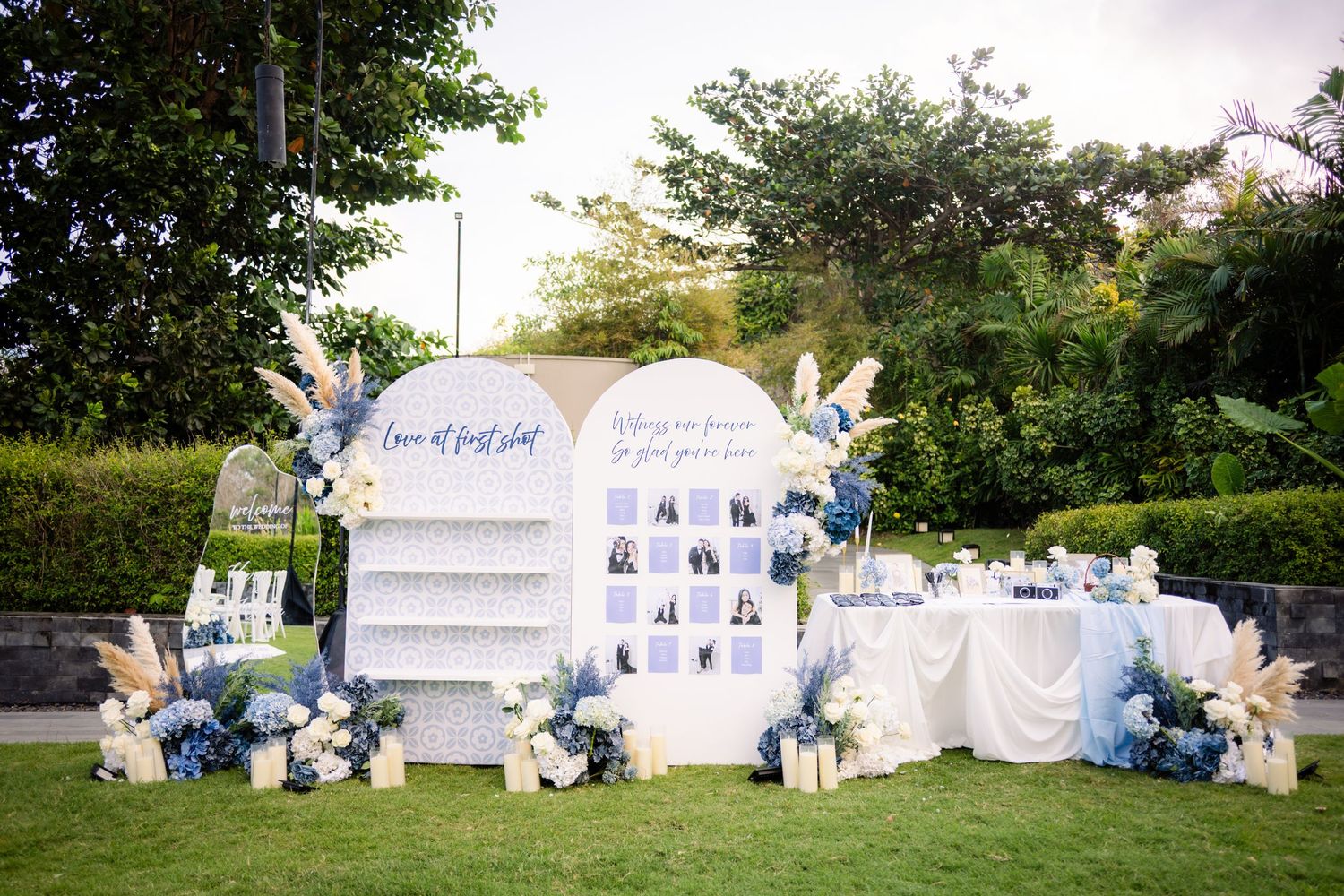 Outdoor garden wedding reception setup with a blue and white seating chart display, floral arrangements, and a draped table on a lawn.
