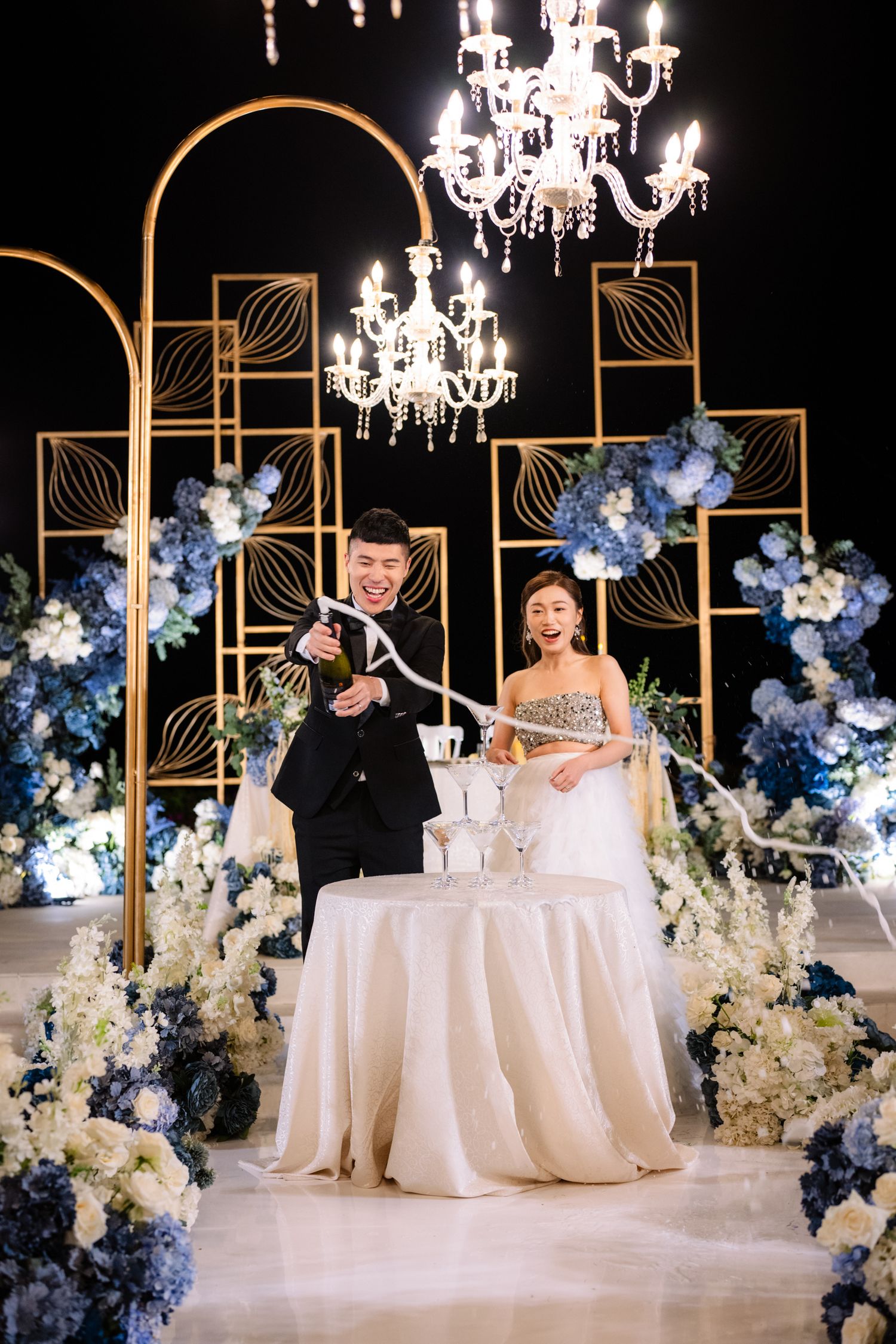 Bride and groom smile beside a draped table as he pops champagne, surrounded by blue and white florals, gold frames and crystal chandeliers.