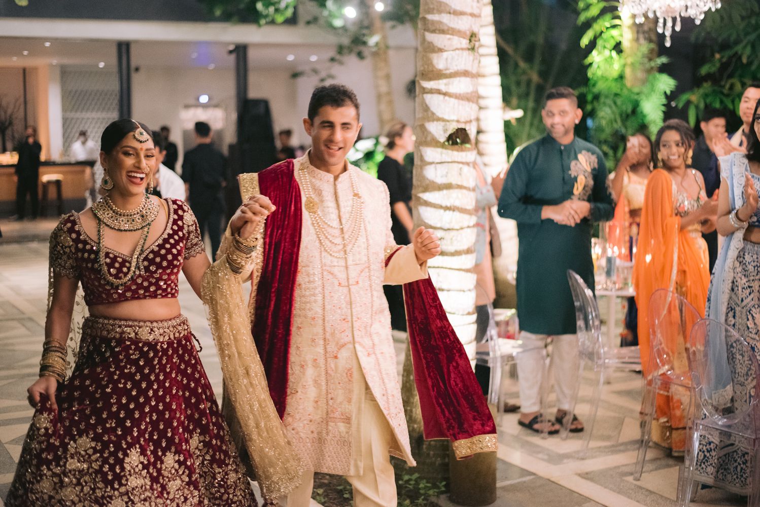 Indian bride in red lehenga and groom in sherwani smiling during wedding ceremony entrance.