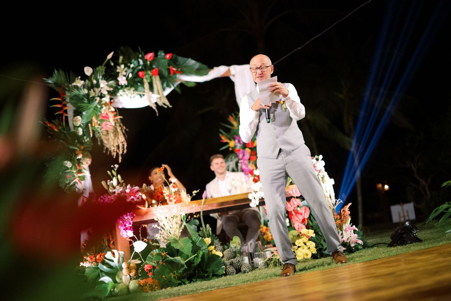 Man in gray suit speaking at decorated outdoor wedding ceremony stage at night.