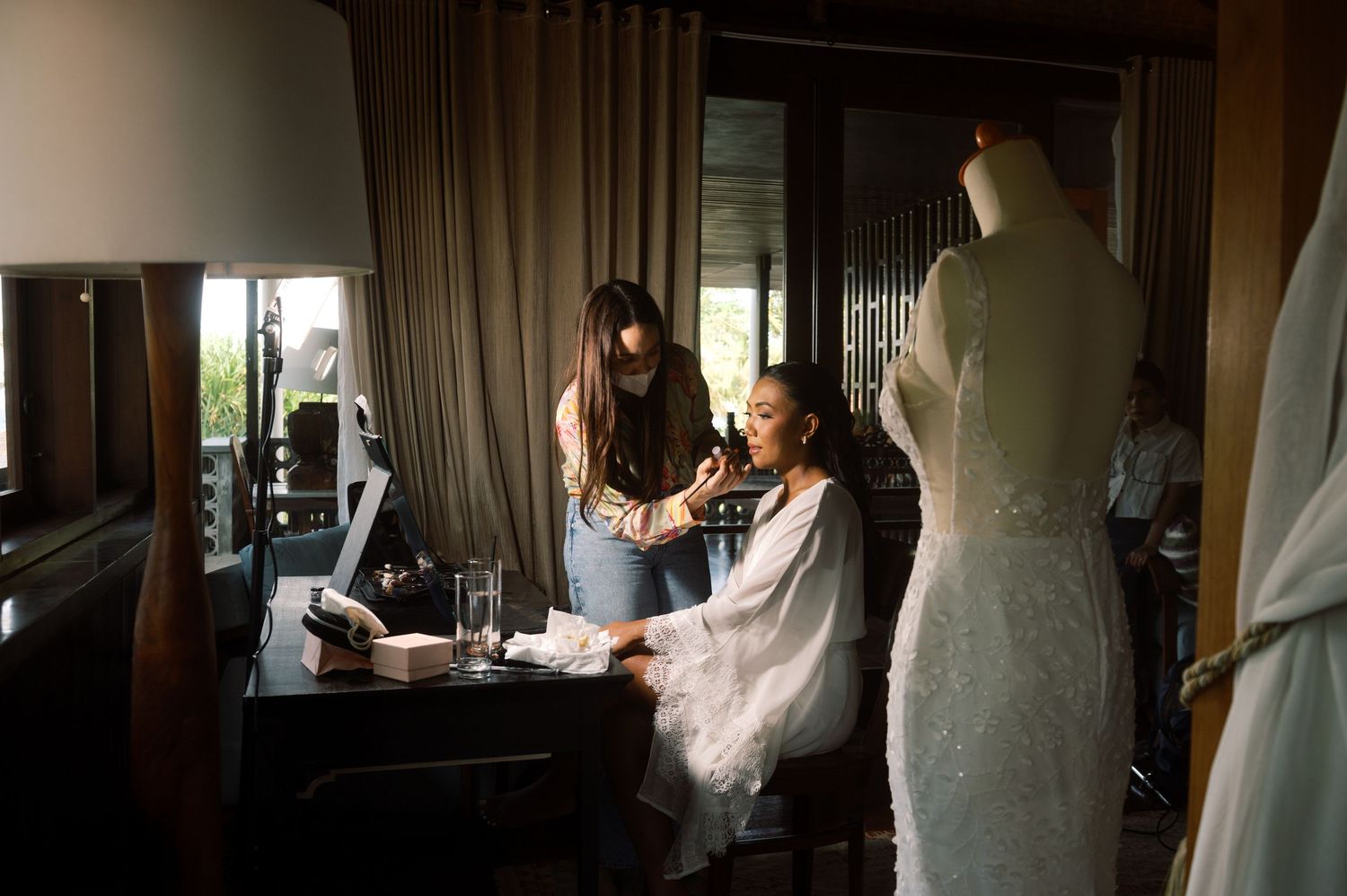 Bride getting makeup done by artist in elegant room with wedding dress on mannequin in background.