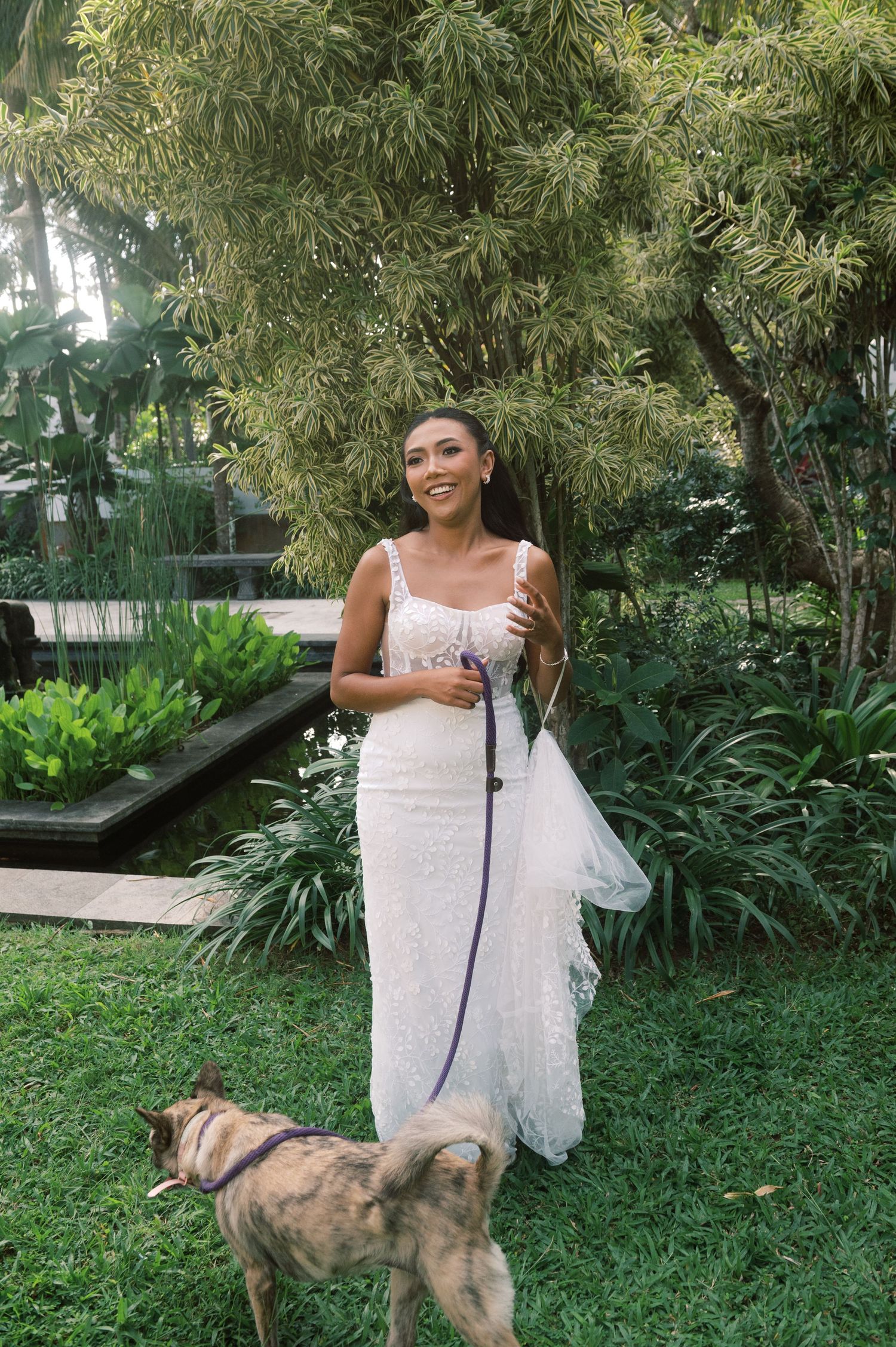 Smiling bride in white lace dress walks dog on leash in lush tropical garden setting.