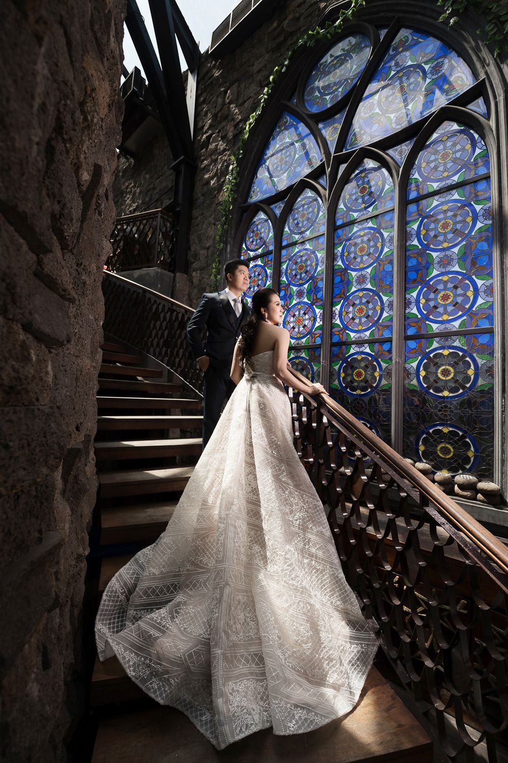 Bride in elegant lace gown and groom on ornate staircase before stunning stained glass window in castle setting.