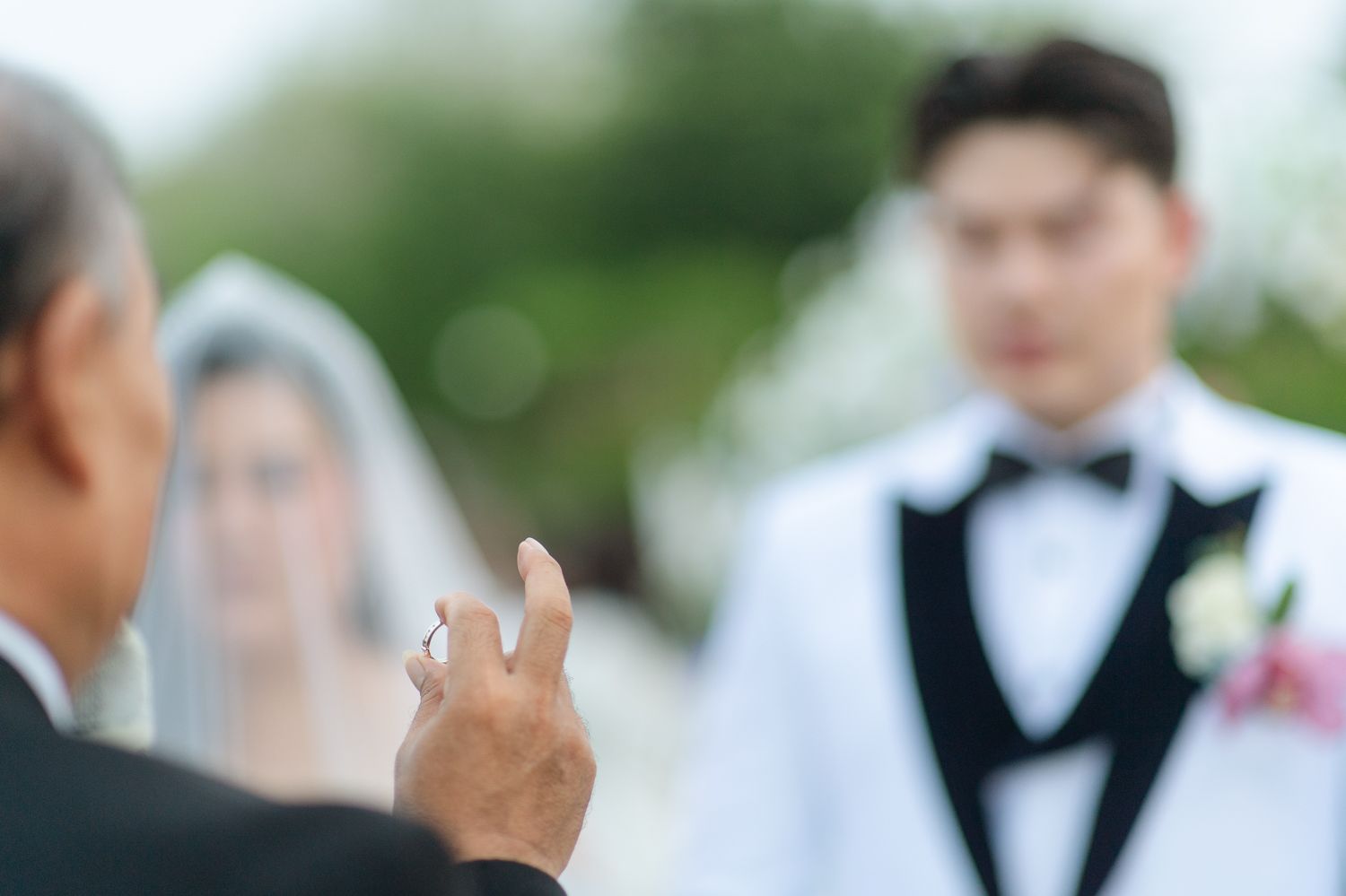 Officiant holds wedding ring during outdoor ceremony with bride in veil and groom in tuxedo in background.
