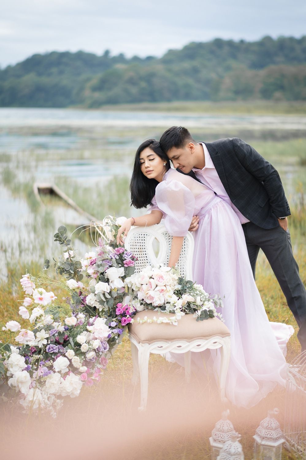 Couple in elegant attire poses romantically near floral arrangement by a serene lake during an outdoor engagement shoot.
