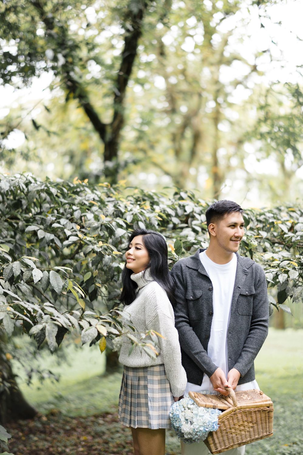 A young couple walks through a lush green park, the man carrying a wicker picnic basket with blue flowers.