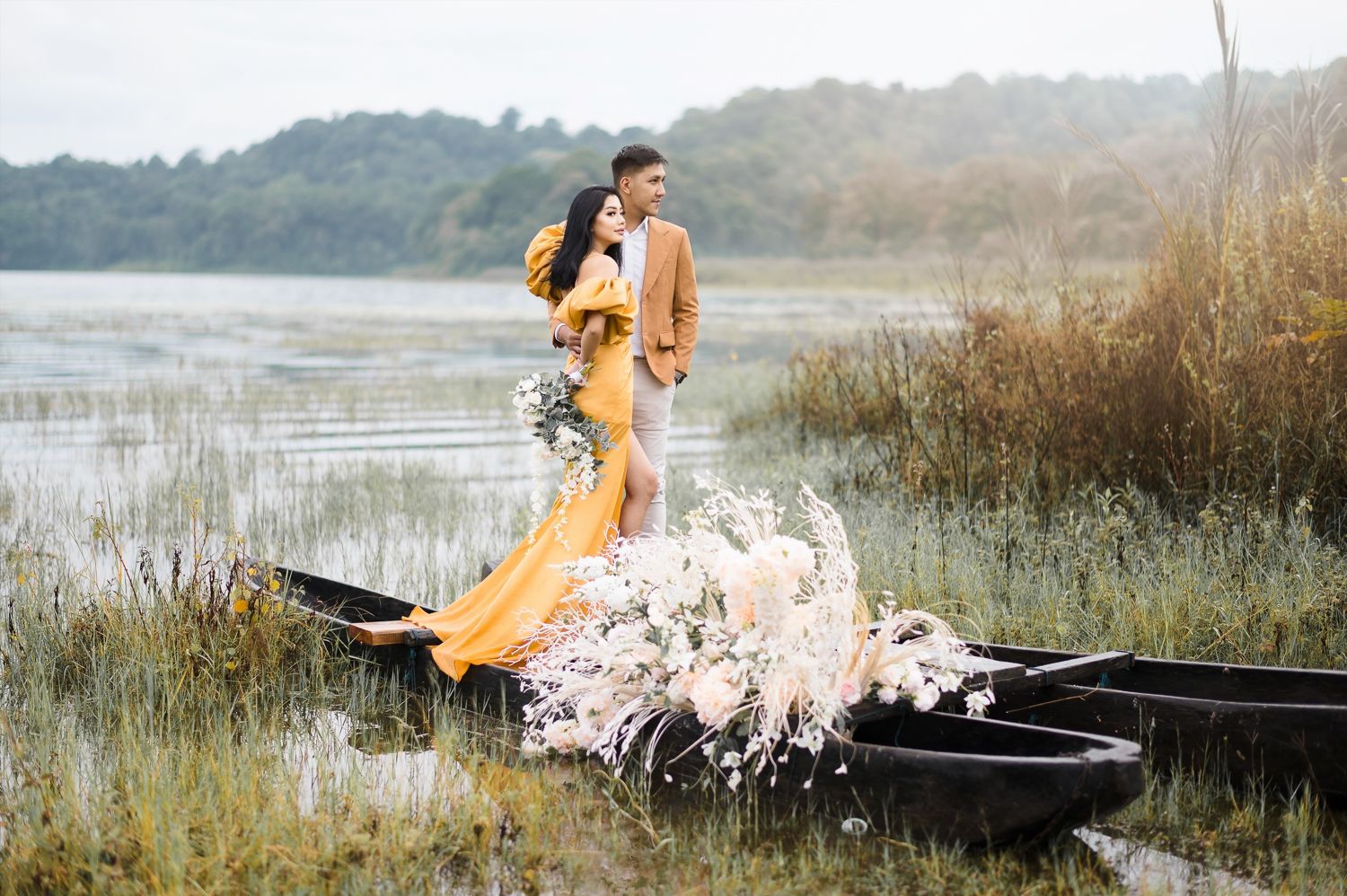 Couple in romantic boat photoshoot, woman in yellow gown, man in tan suit, surrounded by white flowers on a serene lake.