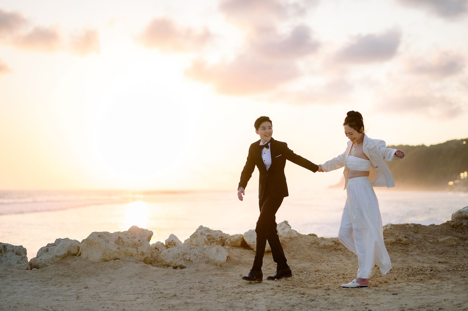 Couple in wedding attire holding hands on a sandy beach at sunset with ocean waves in the background.