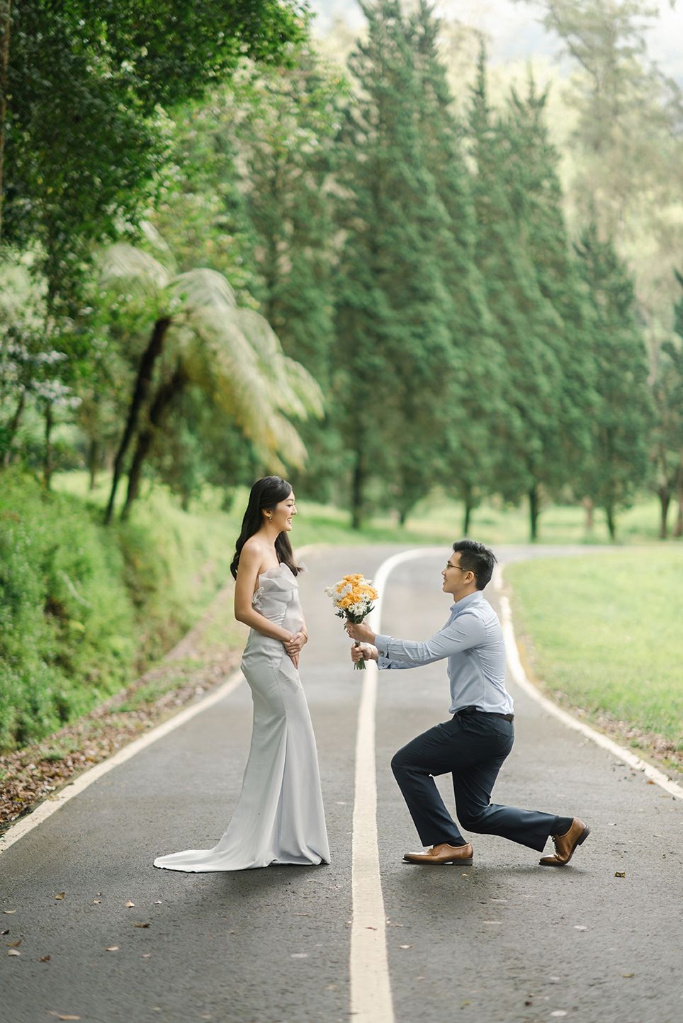 Man kneeling on one knee offering flowers to woman in elegant gown on a tree-lined road in a park.