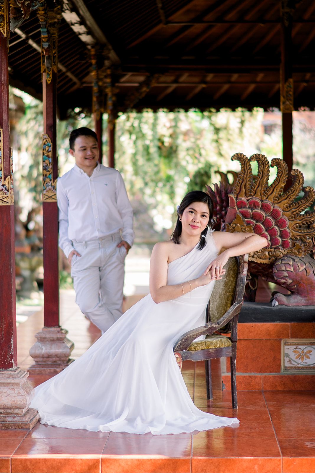 Couple in white attire poses at a Balinese temple pavilion with ornate wooden carvings and red pillars.