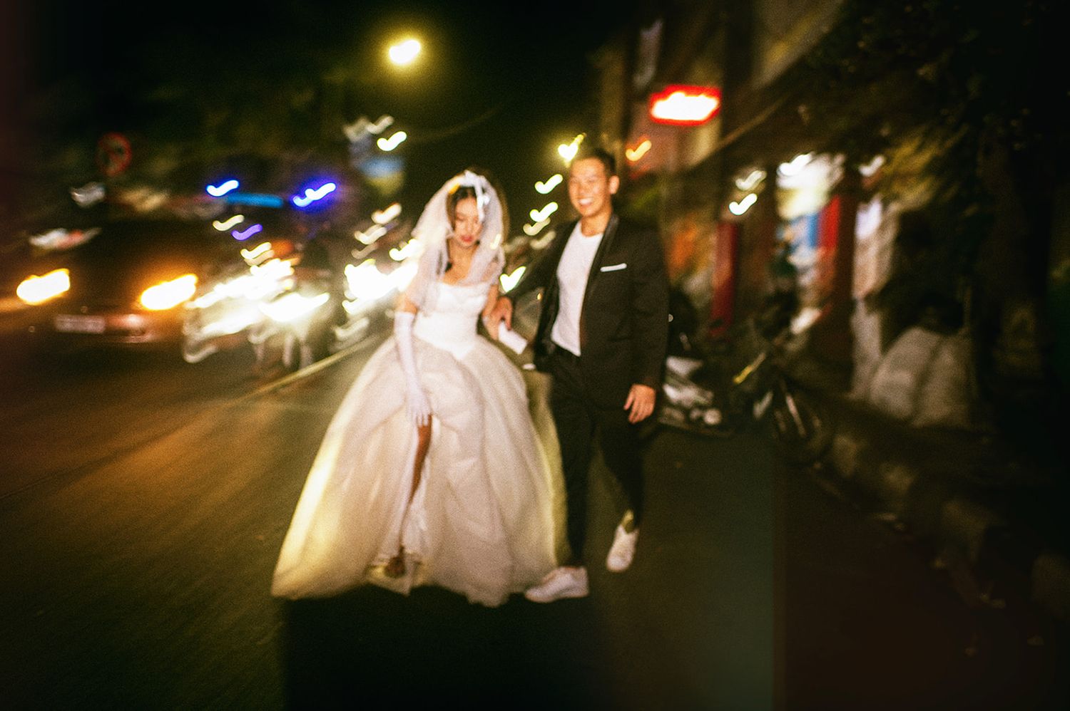 A bride in a white gown and groom in a black suit walk together on a city street at night with bokeh lights.