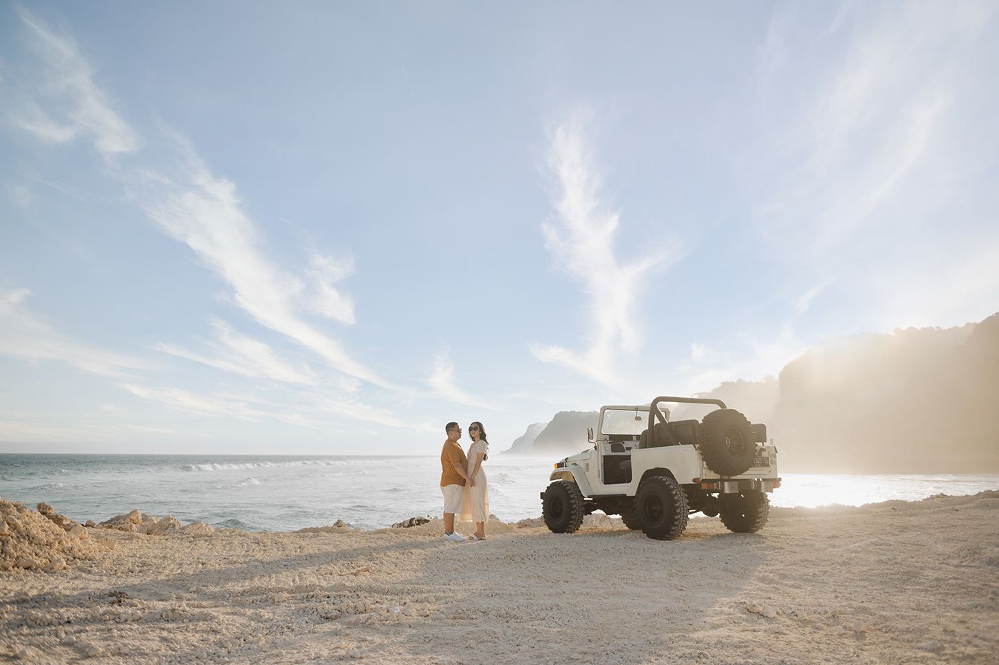 Couple standing together on a sandy beach beside a white vintage Jeep with ocean waves and misty cliffs in the background.