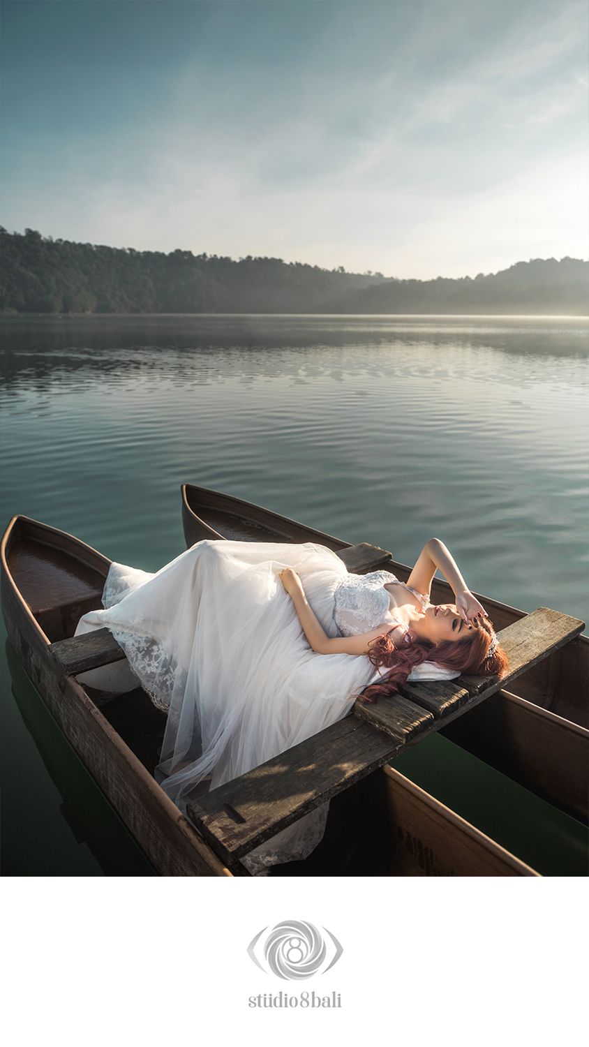 Woman in white wedding dress lying gracefully in a wooden boat on a serene lake at sunrise.