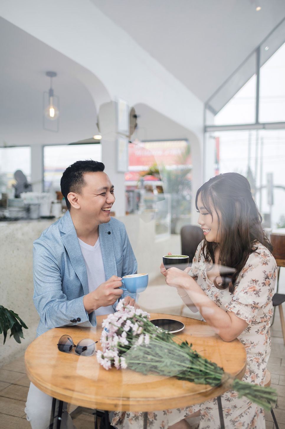 A smiling Asian couple enjoys coffee together at a wooden cafe table with fresh flowers, dressed in smart casual attire.