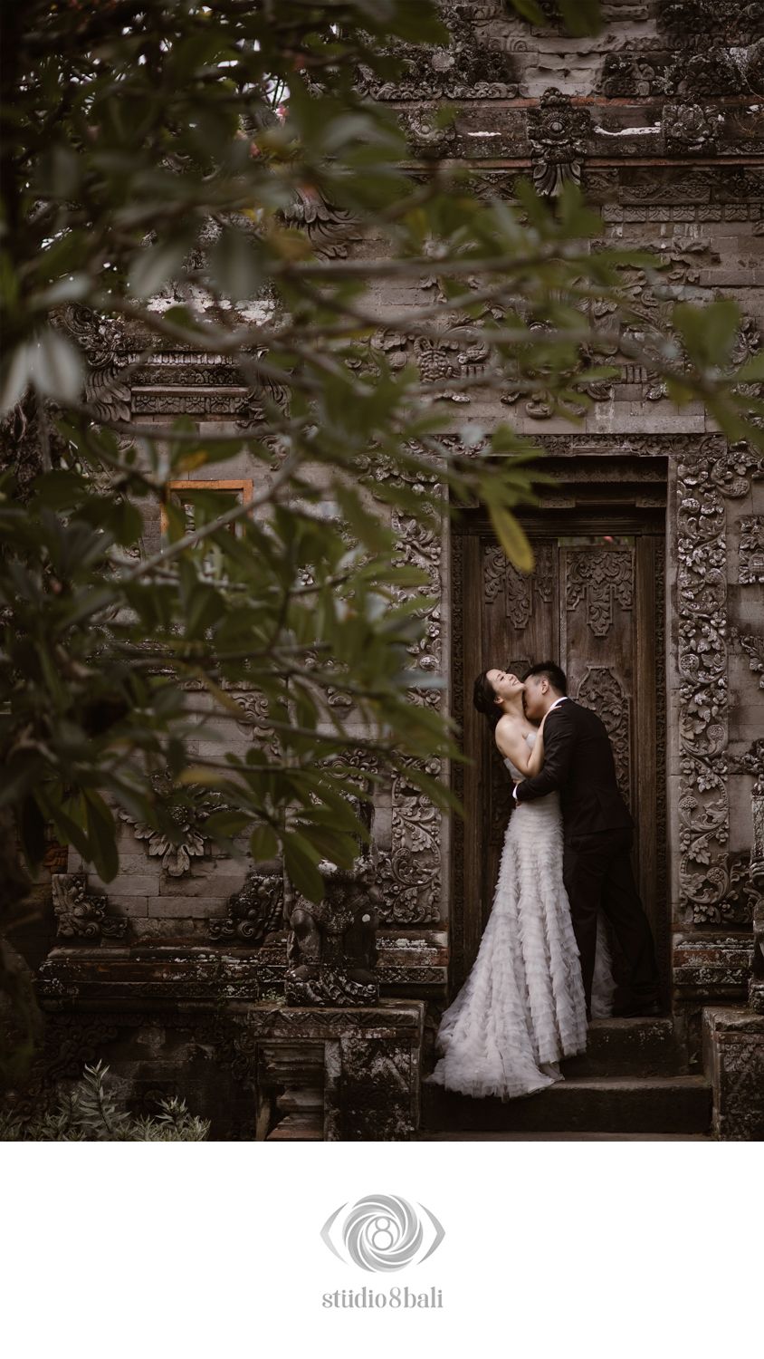 Couple embracing in front of ornate Balinese temple door, bride in lace gown, groom in black suit, framed by lush greenery.