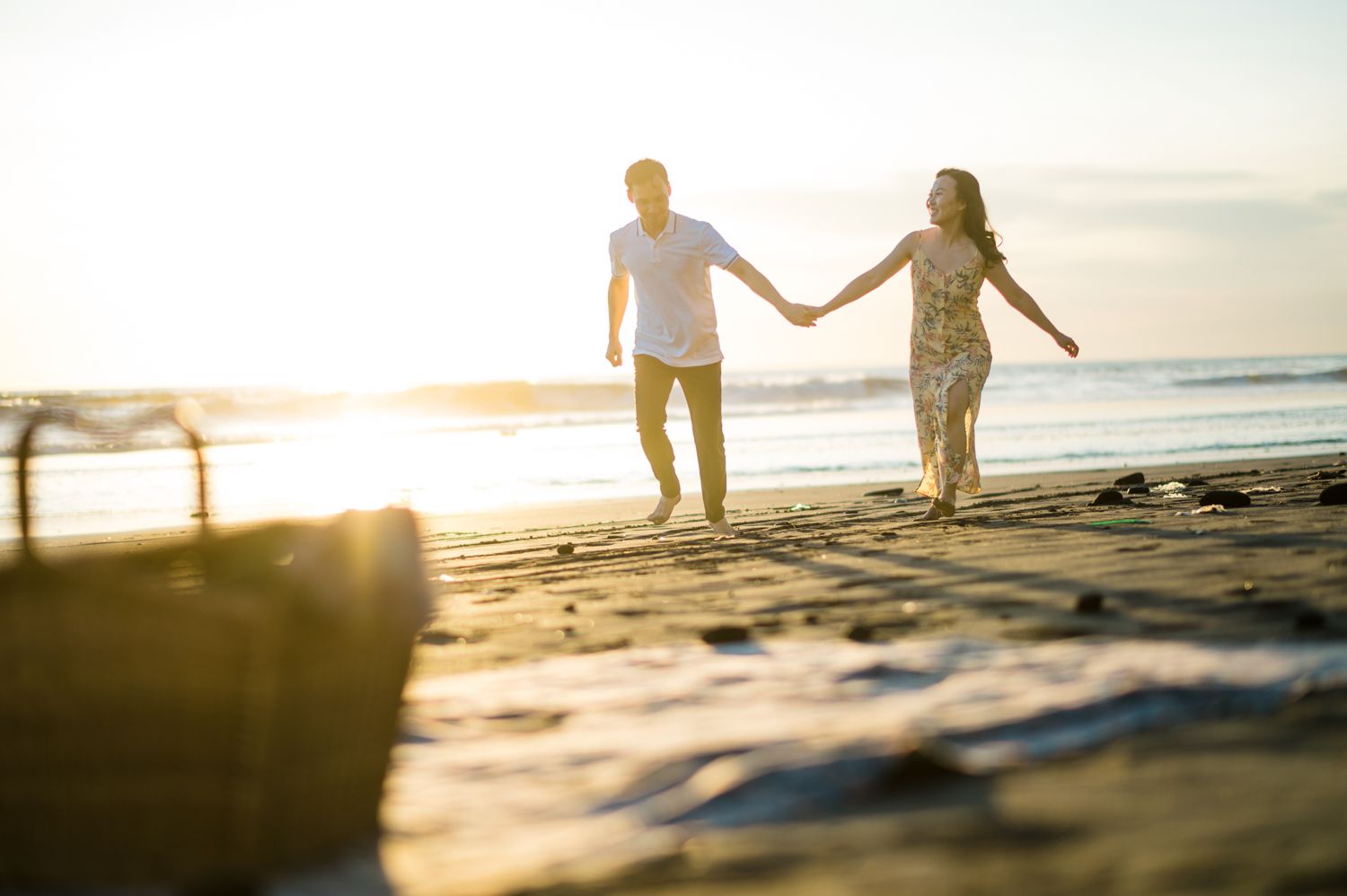 A couple holds hands while walking on a sunny beach at golden hour with a picnic basket in the foreground.