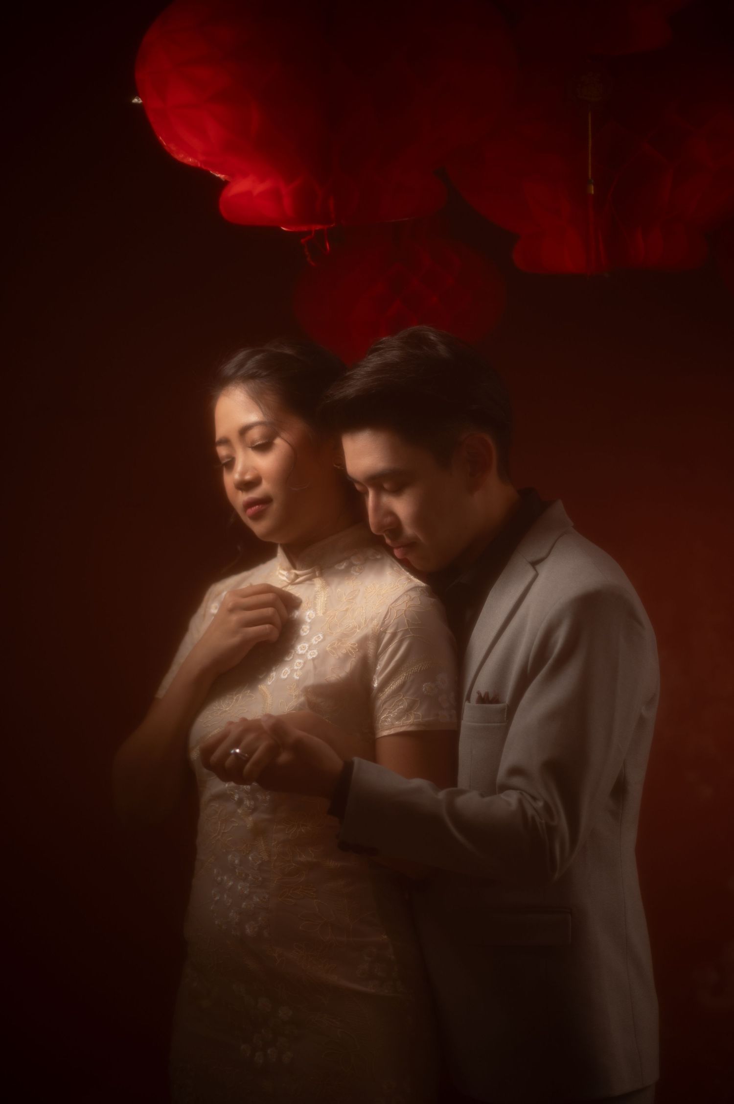 A couple in elegant attire poses intimately under red floral decor in a moody, romantic portrait.
