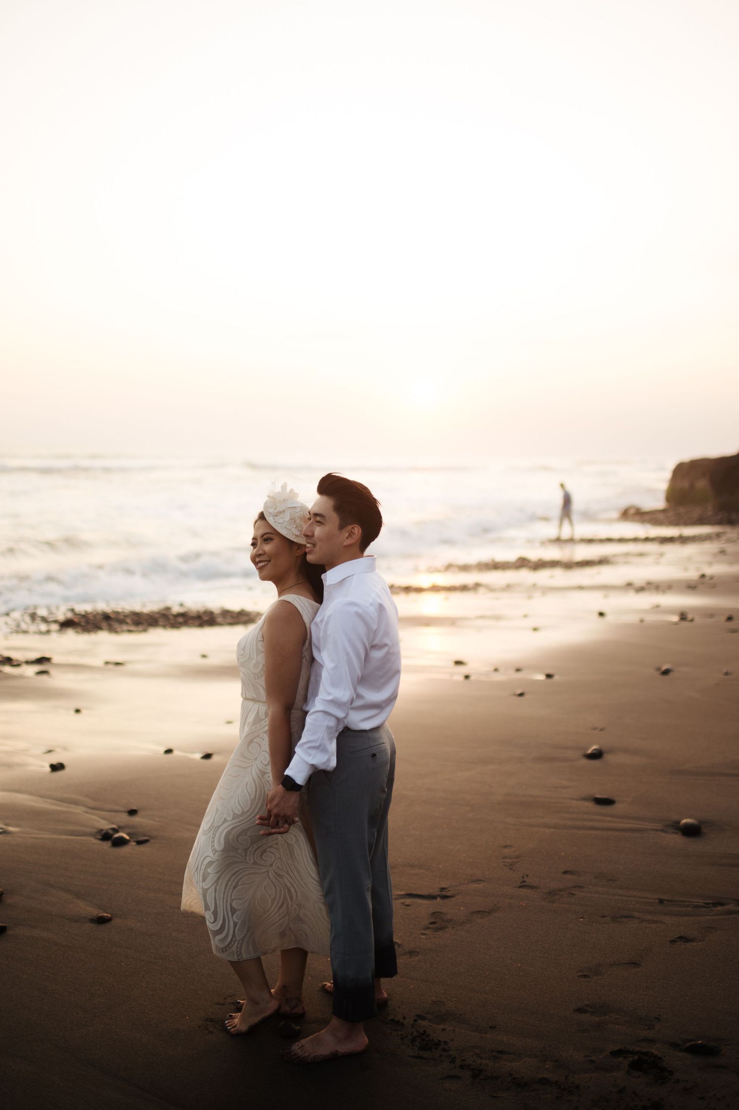 Couple in wedding attire embracing on a sandy beach at sunset with ocean waves in the background.
