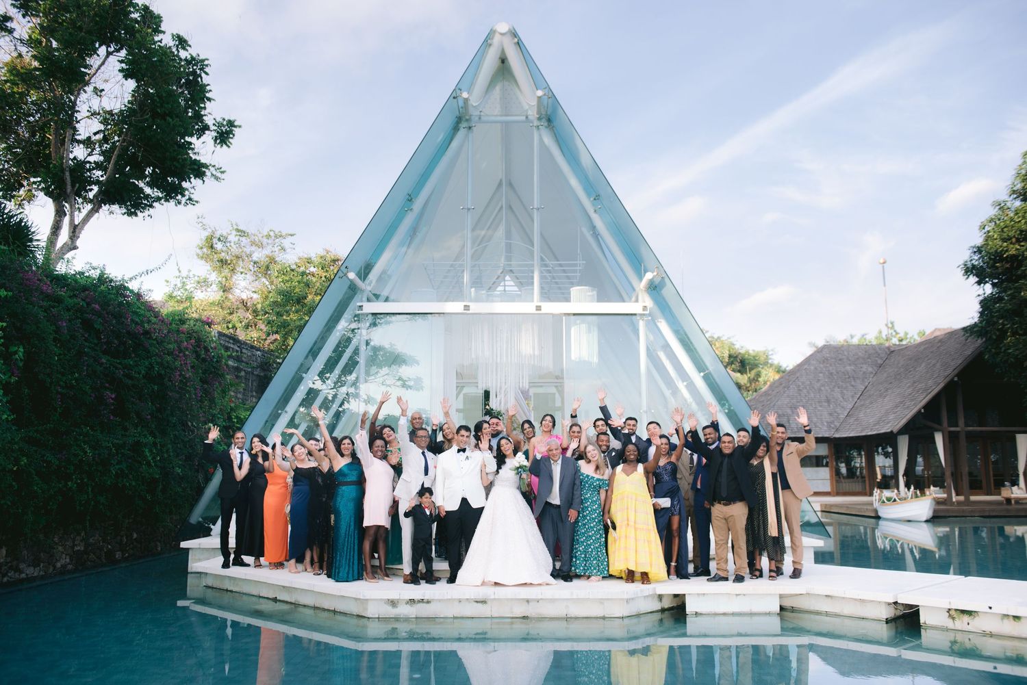 Wedding party celebrating in front of a stunning glass A-frame chapel surrounded by a reflective pool at a tropical venue.
