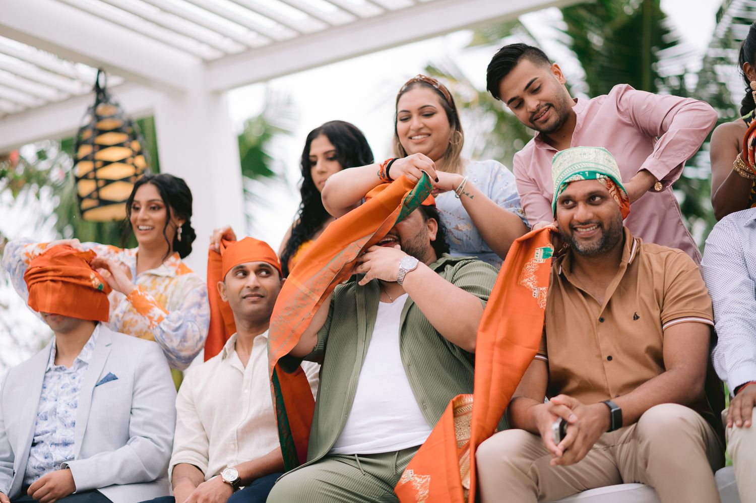 Guests wrap an orange turban on a smiling man during a festive Indian wedding celebration outdoors.