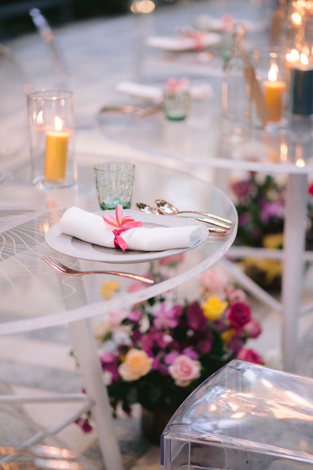 Elegant wedding table setting with white napkins, pink ribbon, candles, and colorful floral centerpieces on a glass table.