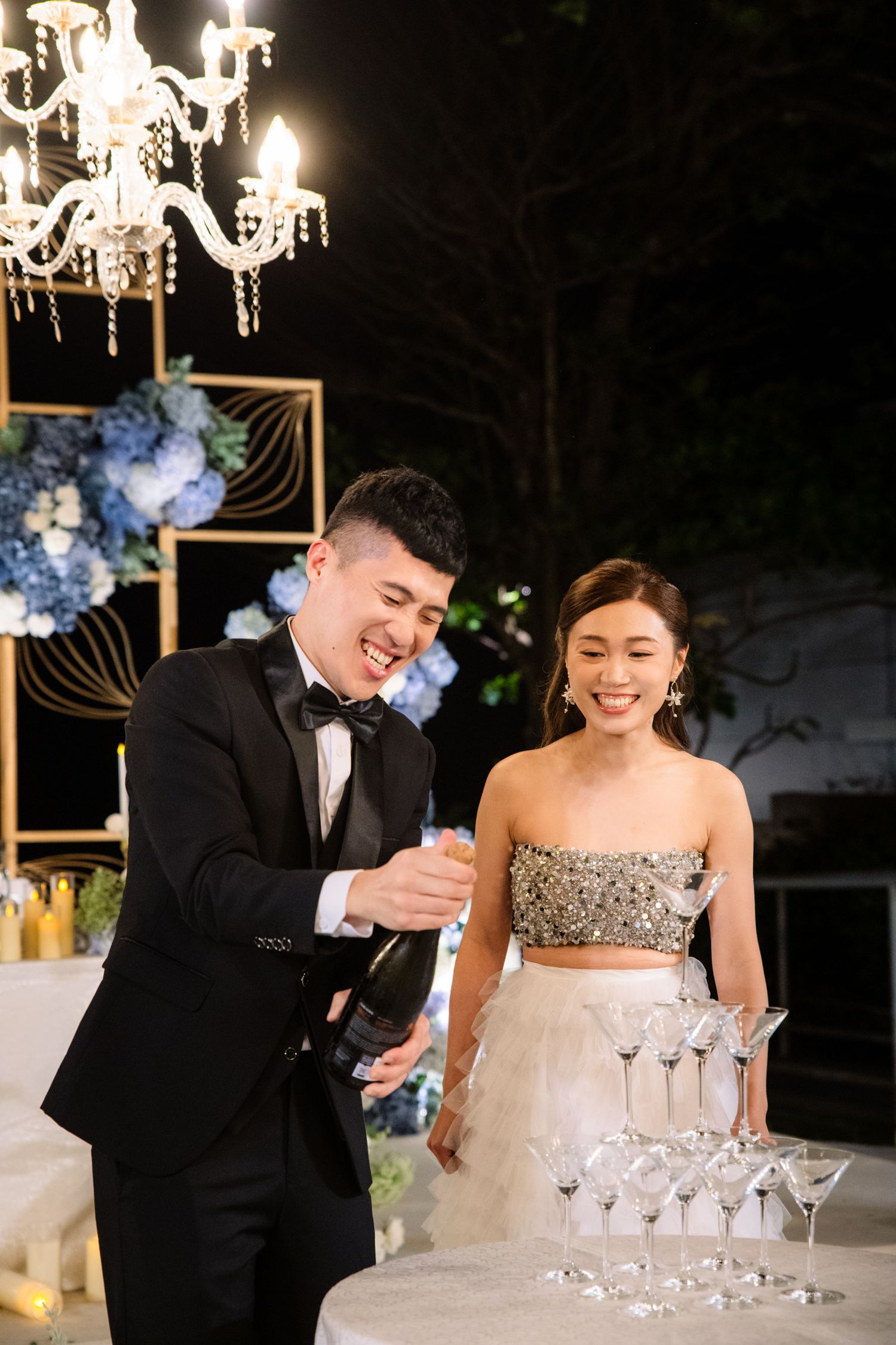 Smiling groom in a black tuxedo pours champagne into a glass tower while his bride in a sparkling crop top laughs beside him at a wedding.