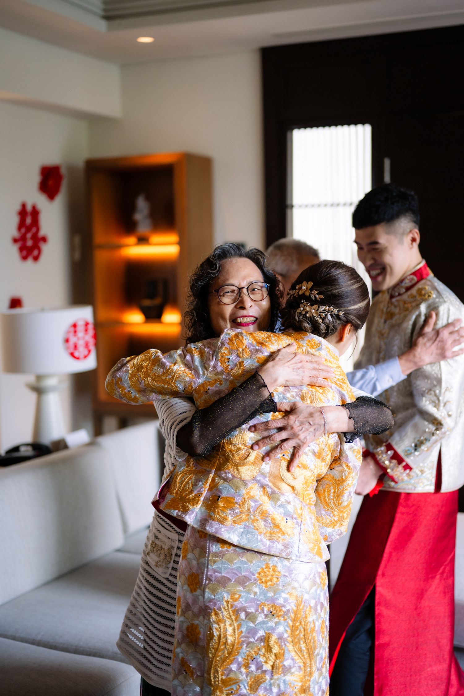 Bride in an embroidered gold Chinese wedding gown hugs a smiling older woman while the groom in traditional attire looks on.