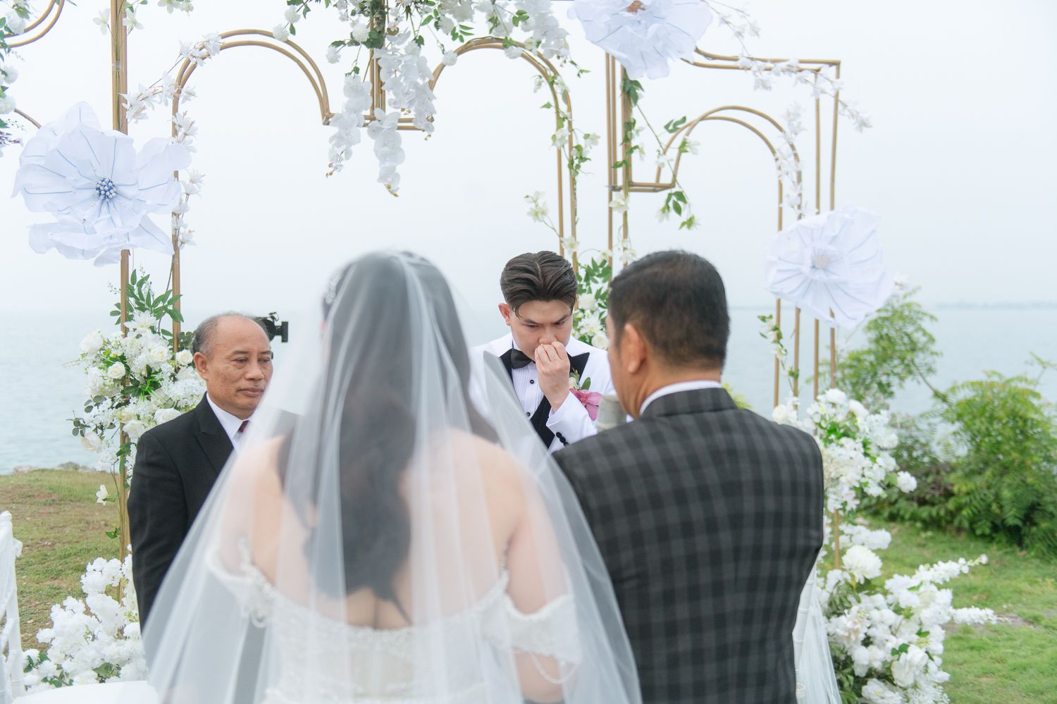 Outdoor wedding ceremony by the water with bride in veil, groom in suit, and guests gathered near white floral decorations.