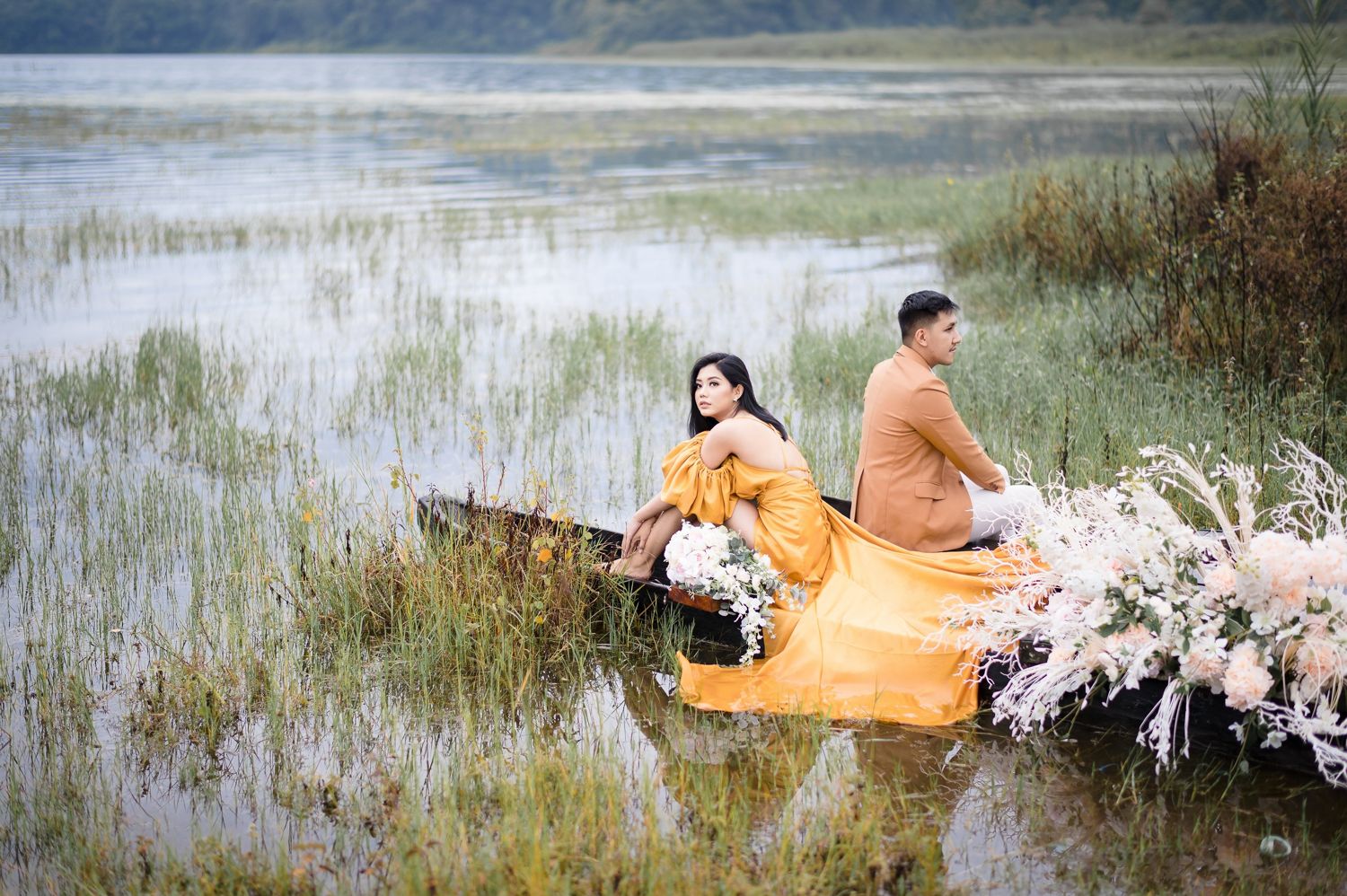 Couple in yellow and orange outfits sitting in a flower-adorned boat on a serene lake surrounded by tall grass.